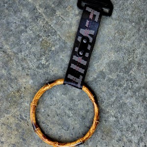 May include: A circular bamboo handle attached to a black nylon strap. The strap features a repeating white and red graphic pattern and is secured to a black metal mounting bracket at the top. The bamboo ring has a natural wood grain finish.