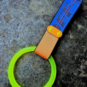 May include: A neon green ring attached to an orange and blue strap. The blue strap has black text and pink stitching. The strap is connected to the ring with an orange piece and yellow fasteners. The item is on a gray surface.