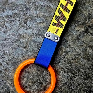 Puede incluir: Una correa amarilla y negra con la palabra "HKH" impresa, conectada a una correa azul y un anillo naranja. La correa está sujeta con herrajes metálicos plateados. El fondo es una superficie gris texturizada.