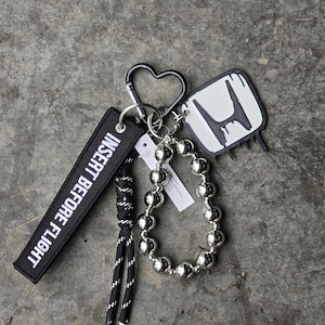 Op de afbeelding: Een verzameling sleutelhangers en accessoires. Inclusief een zwarte hartvormige karabijnhaak, een zwart-wit "INSERT BEFORE FLIGHT" label, een zilveren kralenarmband en een Honda logo sleutelhanger. De items zijn gerangschikt op een grijze, getextureerde ondergrond.