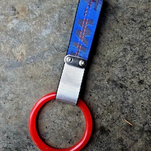Puede incluir: Una correa azul con un patrón negro, asegurada con un elemento metálico plateado y una correa blanca, conectada a un asa circular roja. El patrón de la correa presenta formas negras y líneas rosas.