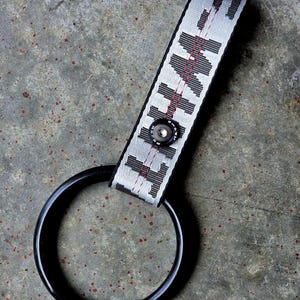 May include: A black circular ring attached to a light grey fabric strap featuring the black text "FLIGHT" and red stitching. A small black metal snap button is centred on the strap, securing the loop that holds the ring in place.