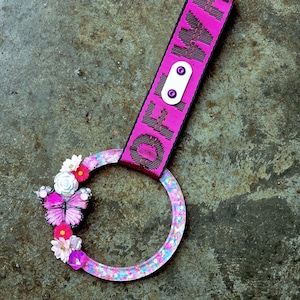 Puede incluir: Un llavero rosa y negro con un diseño circular lleno de purpurina. El llavero presenta una correa rosa con la palabra "OFF" impresa en negro. Un adorno de mariposa y flores adorna la parte circular.