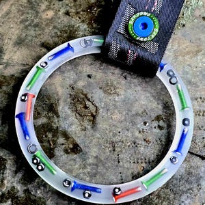 May include: A clear, circular object with embedded colorful bolts and nuts. The bolts are red, green, and blue. A black strap with a circular, blue and green button is attached to the object.