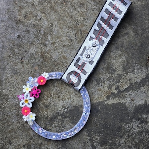 Puede incluir: Un anillo transparente lleno de purpurina con un adorno floral y una correa en blanco y negro que dice "OFF WHITE". El anillo presenta flores rosas, blancas y azules. La correa está sujeta al anillo con un cierre de metal plateado.