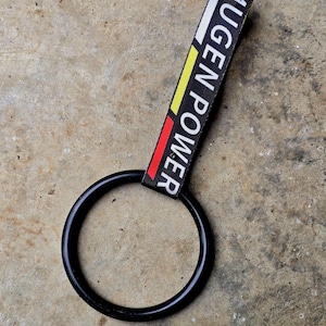 Puede incluir: Llavero negro con una correa negra, blanca, roja y amarilla. La correa dice "MUGEN POWER" en texto blanco. El llavero es circular y está hecho de un material negro brillante.
