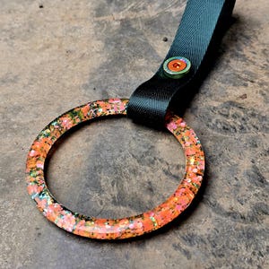 Puede incluir: Un llavero decorativo con un anillo naranja brillante con detalles en rosa y verde. El anillo está unido a una correa de tela verde oscuro asegurada con un botón. El llavero está diseñado para guardar llaves.