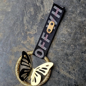 Puede incluir: Un anillo dorado con purpurina y diseño de mariposa. La mariposa tiene un ala negra y otra dorada. Una correa negra con la palabra "OFF" en blanco y rosa está unida al anillo.
