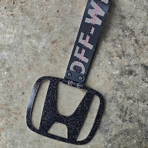 Puede incluir: Llavero negro con el logotipo de Honda y una correa en blanco y negro que dice "OFF WHITE" en letras mayúsculas blancas con una raya rosa. El llavero está sobre una superficie de hormigón gris.