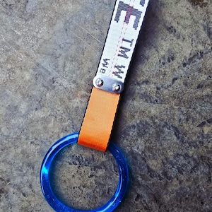 Puede incluir: Un anillo azul translúcido unido a una correa naranja y una correa blanca con texto negro. La correa blanca tiene el texto "TM W" y "WE" impreso. El anillo está hecho de un material transparente y brillante.
