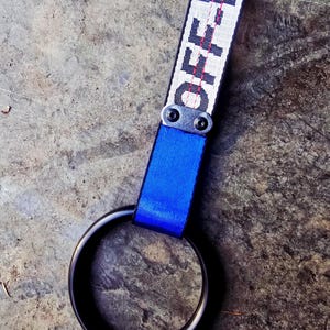 Puede incluir: Un anillo negro unido a una correa azul y una correa blanca con la palabra "OFF WHITE" impresa en negro. Las correas están conectadas con una pieza de metal. El fondo es una superficie gris texturizada.
