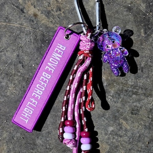 Op de afbeelding: Een paarse sleutelhanger met een zilveren karabijnhaak. De sleutelhanger heeft een paars label met "REMOVE BEFORE FLIGHT" in witte tekst, een gevlochten kwast met roze en rode accenten en een kleine paarse berenbedel met glitter.