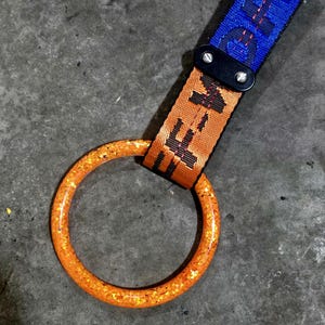 May include: Close-up of a key ring with a vibrant orange glittery ring. The strap is blue with white text and orange with black text. The key ring is attached with a black leather piece.