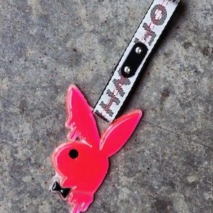 May include: A neon pink translucent rabbit head pendant with a black eye and bowtie, attached to a white woven strap featuring black text and red stitching. The strap includes a black leather-like tab secured with two silver-toned metal rivets.