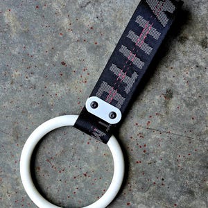 May include: A white circular handle attached to a black nylon strap. The strap features repeating white text and thin red stitching. A white rectangular plastic piece with two black rivets secures the strap to the handle, creating a loop design.