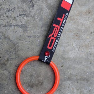 Puede incluir: Una correa negra TRD Racing Development con un anillo rojo y naranja. La correa tiene texto blanco y un diseño rectangular rojo y blanco. Una joya roja está unida a la correa.
