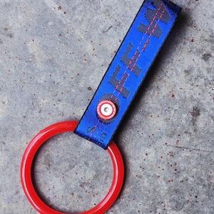 May include: A blue nylon strap keychain featuring black text and a thin red stitched line, secured with a silver and red circular metal rivet. A solid red plastic ring is attached to the bottom of the strap, providing a functional loop for keys.