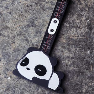 Puede incluir: Una etiqueta de equipaje con forma de panda en blanco y negro unida a una correa de tela negra. La correa presenta texto en rojo y blanco y está asegurada con un cierre de plástico blanco que contiene dos remaches negros. El diseño es plano.
