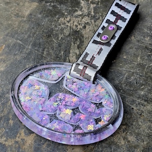 Puede incluir: Llavero ovalado de resina transparente con un diseño en forma de estrella lleno de purpurina morada e iridiscente. El llavero tiene una correa blanca y negra con la palabra "F***" impresa.