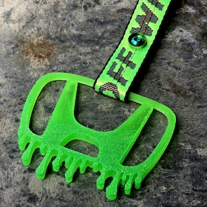Puede incluir: Un llavero verde neón con un logotipo modificado y un efecto de goteo. El llavero está unido a una correa estampada verde y negra con la palabra "HESH" y un botón decorativo.