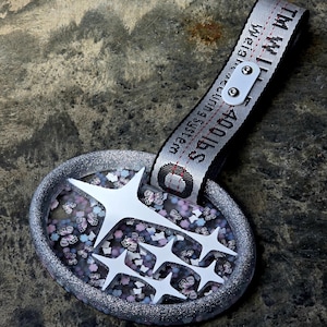 Puede incluir: Un accesorio ovalado con un borde plateado brillante, con un diseño de estrella blanca y un fondo de mariposas. Una correa blanca con el texto "Weight Securing System 400 lbs" está adjunta.