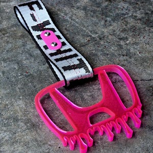 Op de afbeelding: Een neonroze doorschijnende kunststof handgreep in de vorm van een autologo met een druipeffect aan de onderkant. Het is bevestigd aan een witte geweven stoffen band met zwarte tekst en een klein roze rechthoekig lipje vastgezet met twee klinknagels.