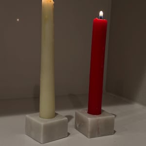 May include: Two lit taper candles, one cream and one red, stand in square marble holders. The cream candle has a tall, slender shape, while the red candle is a similar size. The marble holders are a light gray color.