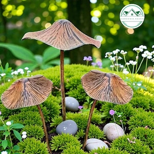Peut inclure: Trois piquets de jardin décoratifs en forme de champignon avec des chapeaux et des tiges bruns sont plantés dans un lit de mousse verte. De petites pierres grises et de minuscules fleurs blanches sont dispersées autour des champignons. Le logo Gardenia Haven est en arrière-plan.
