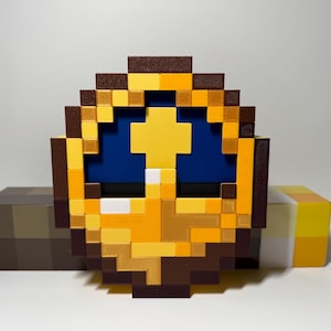 Puede incluir: Un diseño pixelado en 3D, en tonos marrones, amarillos y azules, que se asemeja a una cara estilizada. La obra de arte es en bloques, con una forma de cruz en el centro. Una paleta de colores de bloques marrones y amarillos está en el fondo.