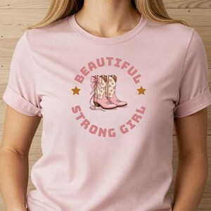 Könnte beinhalten: Hellrosa T-Shirt mit einem kreisförmigen Design. Das Design zeigt die Worte "BEAUTIFUL STRONG GIRL" und zwei goldene Sterne. Das Design beinhaltet ein Paar rosa Cowboystiefel mit floralen Akzenten.