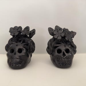 May include: Two black decorative skull figurines. Each skull has a floral crown and detailed carvings. The skulls have large eye sockets and a detailed mouth. The figurines are displayed on a white surface.