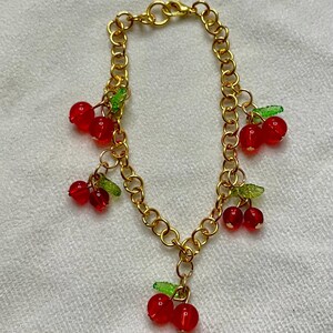 May include: Gold-colored chain bracelet with red cherry charms and green leaf accents. The bracelet features multiple dangling cherry clusters, each with two or three red beads and small green leaves. The chain has a lobster clasp.