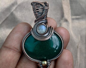 May include: Handmade pendant featuring a polished, oval-shaped green stone. The stone is encased in intricately woven copper wire, with a small, round, light-colored stone accent. The pendant has a loop at the top for a chain.