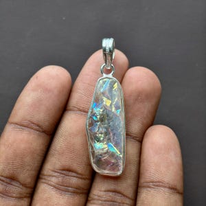 May include: A silver-toned pendant featuring an elongated, irregular-shaped stone with a translucent, iridescent surface. The stone displays internal fractures that reflect flashes of blue, yellow, and green light. It is held by a simple metal bail.