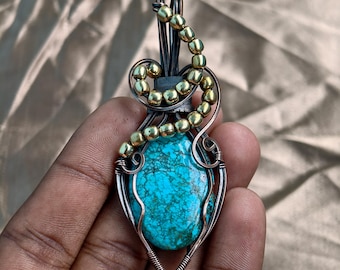 May include: Handmade pendant featuring a vibrant turquoise stone, intricately wire-wrapped with silver and copper wire. Accented with gold-colored beads, the pendant has a unique, artistic design. The turquoise stone is approximately 1.5 inches long.