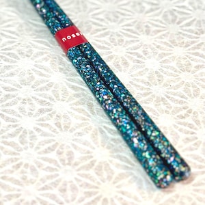 May include: A pair of chopsticks featuring a dark teal and blue iridescent speckled finish with a reflective, glitter-like texture. A small red paper band is wrapped around the upper portion of the chopsticks, displaying the white text "noss".