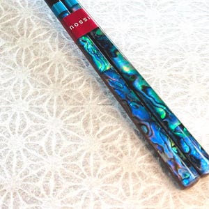 May include: A pair of chopsticks featuring a vibrant iridescent abalone shell inlay with swirling blue and green patterns. The upper portion of the chopsticks is secured by a red band with the white text "nossi" printed on the surface.