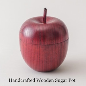 May include: A deep red, apple-shaped wooden sugar pot with a smooth, polished finish and visible wood grain. The container features a small, upright stem on the lid. The text "Handcrafted Wooden Sugar Pot" is printed below the object.