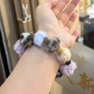 Könnte beinhalten: Ein Armband mit flauschigen Pompons in Rosa, Weiß, Grau und Beige, abwechselnd mit braunen und goldenen Perlen. Ein farbenfrohes Accessoire, das jedem Outfit einen Hauch von Verspieltheit verleiht.