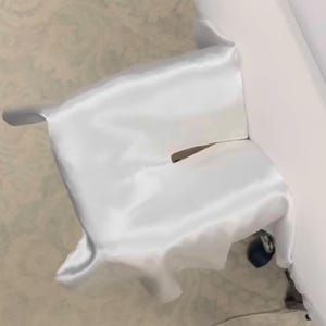 May include: A white satin fabric draped over an unseen object, creating soft folds and shadows. The fabric has a smooth, reflective surface, and a small opening is visible. The overall effect is elegant and minimalist.