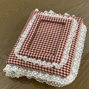 Puede incluir: Un marco de fotos rectangular de tela con un patrón de cuadros vichy en rojo oscuro y crema. El marco está decorado con ribetes de encaje blanco a lo largo de los bordes y alrededor de la abertura rectangular central, creando un aspecto texturizado.