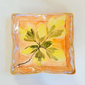 Könnte beinhalten: Eine quadratische Servierplatte aus Keramik mit einem erhöhten Rand und einem bemalten Design eines gelben und grünen Blattes auf einem pfirsichfarbenen Hintergrund.