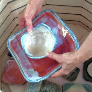 May include: A hand holds a red and blue ceramic dish with a smaller white ceramic bowl inside. The dish has a blue rim and the bowl has a blue rim. The dish and bowl are sitting on a table with other ceramic pieces.
