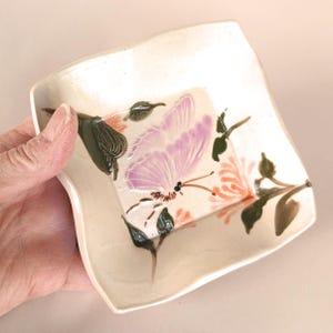 Könnte beinhalten: Eine kleine, weiße Keramikschale mit welligem Rand. Die Schale ist mit einem handgemalten Design eines lilafarbenen Schmetterlings mit orangefarbenen Blumen und grünen Blättern verziert.