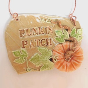 Puede incluir: Un letrero de cerámica con las palabras "PUNK'N PATCH" en letras marrones. El letrero presenta una calabaza naranja en 3D, hojas verdes y una vid retorcida. El letrero es de color beige con un alambre de cobre para colgar.