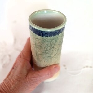 May include: A small, cylindrical ceramic vase with a light green glaze and a blue band around the top. The vase has a textured floral design.