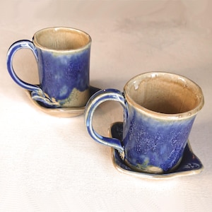 May include: Two handmade ceramic mugs with matching saucers. The mugs are a deep blue with a textured glaze, and the interiors are a natural beige. Each mug sits on a small, organically shaped saucer, creating a rustic and artistic aesthetic.