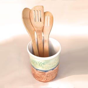 May include: A ceramic utensil holder with a green and blue floral design and a brown textured base. Three wooden spoons are inside the holder.