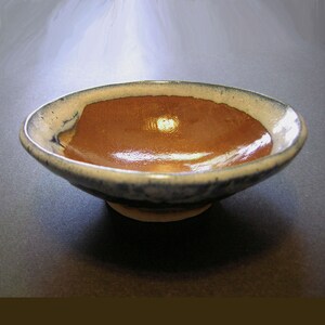 Op de afbeelding: Een kleine, bruine keramische kom met een witte rand en blauwe glazuur. De kom heeft een licht onregelmatig oppervlak en een glanzende afwerking.