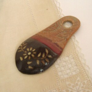 Pode incluir: Uma colher de cerâmica marrom com um esmalte vermelho e bege. A colher tem um desenho floral branco sobre o esmalte marrom. A colher tem um furo na parte superior para pendurar.